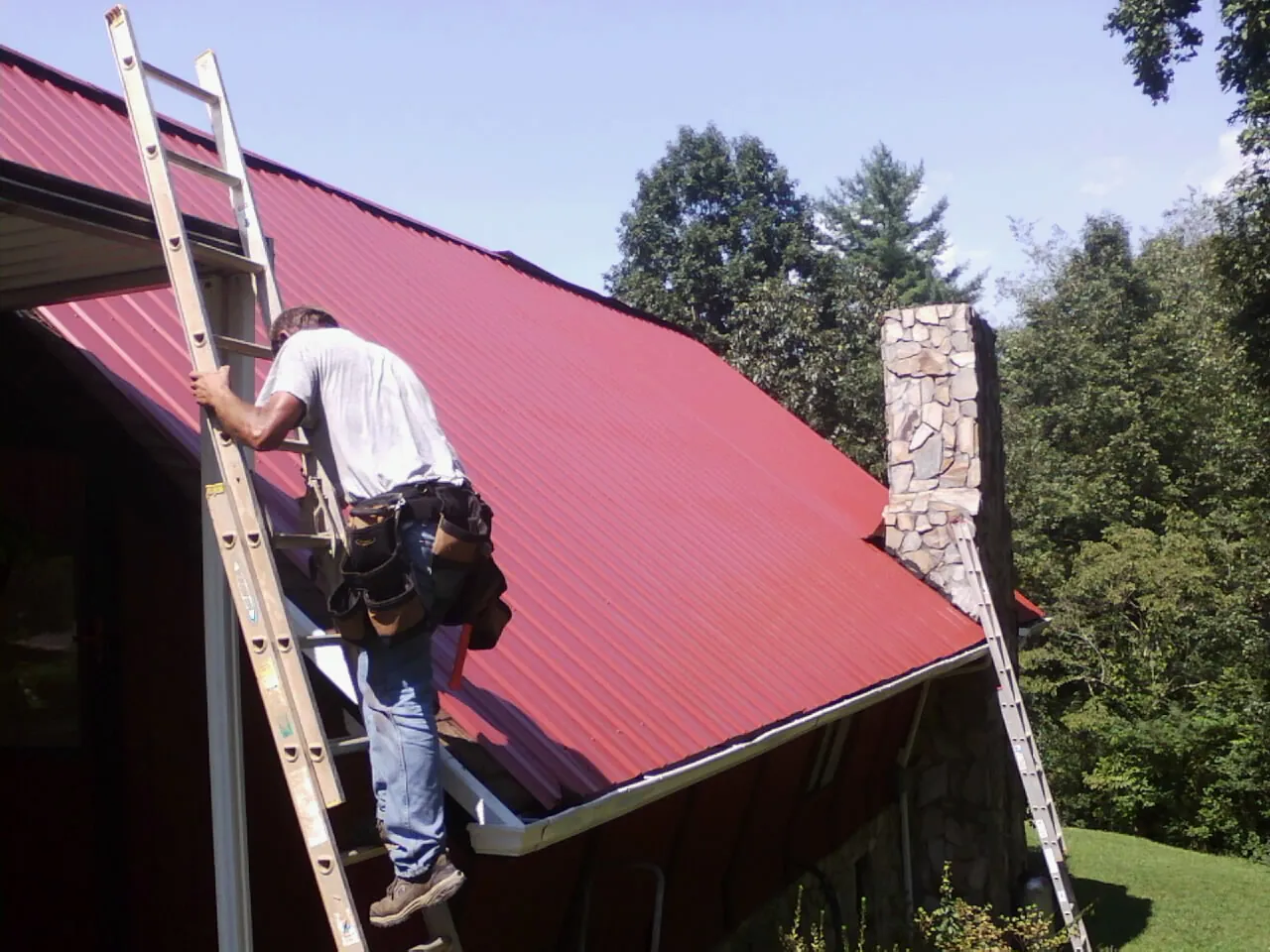 Roofer on a ladder beside a red metal roof and stone chimney — Soffit & Fascia Repair by Roofing Brothers in Pinson